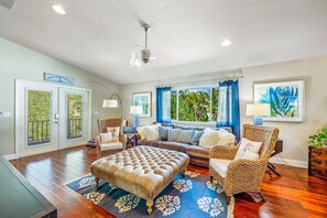 A spacious living room with a large sofa, two armchairs, a coffee table, and a ceiling fan. There are two doors with glass panels, a window with blue curtains, and a framed artwork on the wall. - Decked Out: Steps to Bay & Beach, Pool, Games by Anna Maria Life Vacation Rentals (Anna Maria)