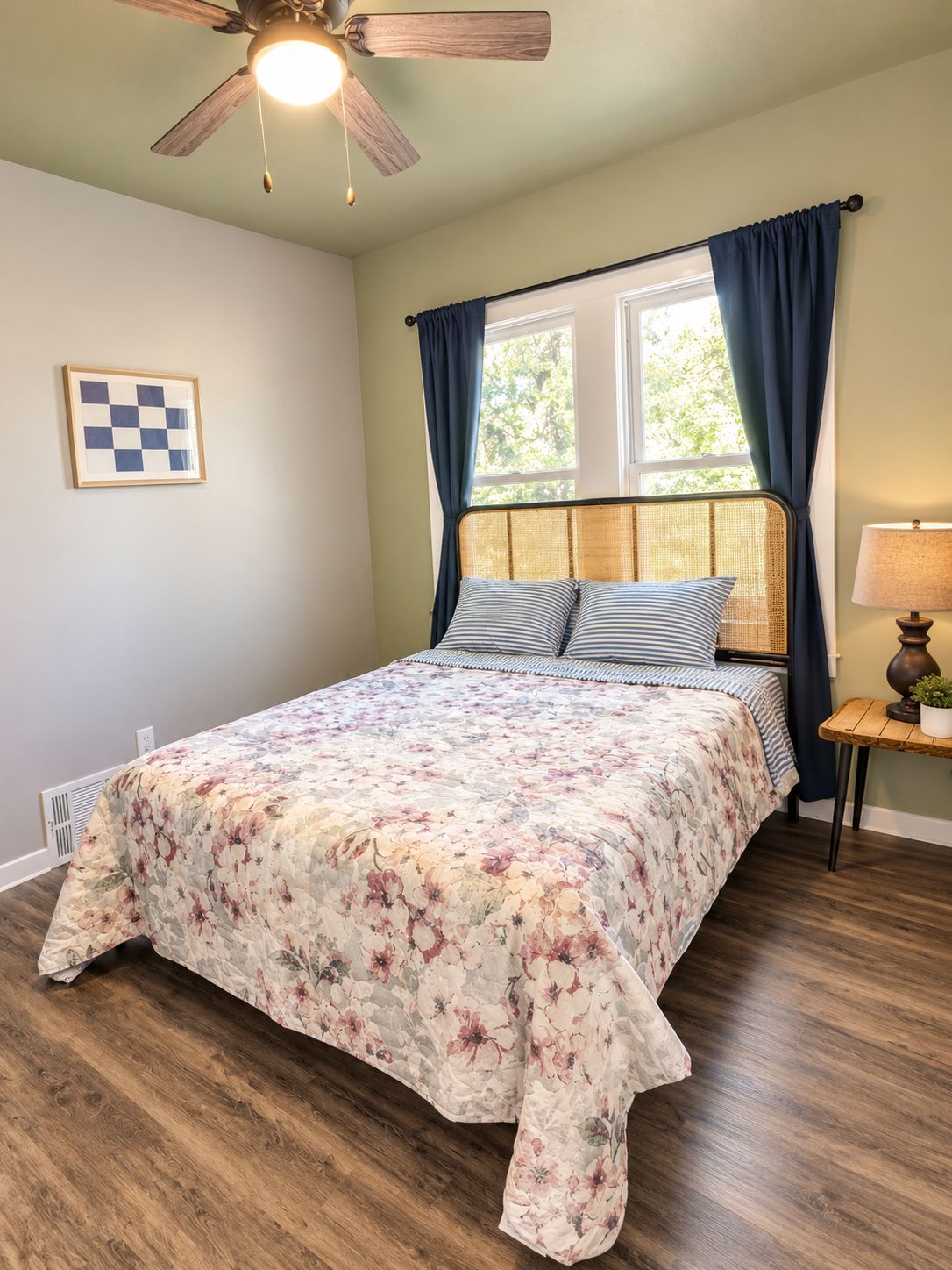 Een slaapkamer met een bed, een plafondventilator, een raam met gordijnen, een nachtkastje met een lamp en een lijst met een afbeelding aan de muur.