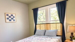 Une chambre à coucher avec un lit, un ventilateur de plafond, une fenêtre avec des rideaux, une table de chevet avec une lampe et un tableau encadré au mur.