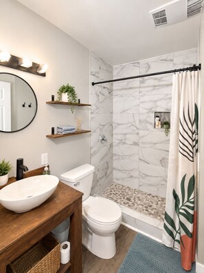 Ein Badezimmer mit Marmordusche, Toilette in Weiß, Holzwaschtisch und einem Vessel-Becken sowie einem Spiegel.