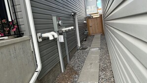 En smal gångväg med en kant av betong, omgiven av grå hus med träpanel, en trädörr och en metallåda för el- eller vattenledningar.