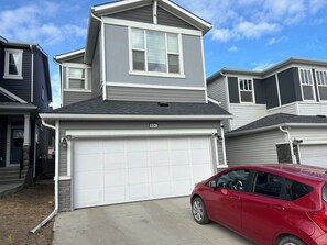 Une maison à deux étages avec un garage, une voiture rouge garée dans l’allée, et un ciel bleu dégagé.
