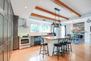 A modern kitchen with wooden beams, a large island with bar stools, stainless steel appliances, and a stone fireplace. - Chalet Petit Bonheur - Waterfront (beautiful beach) - Fully equipped (Val-des-Bois)