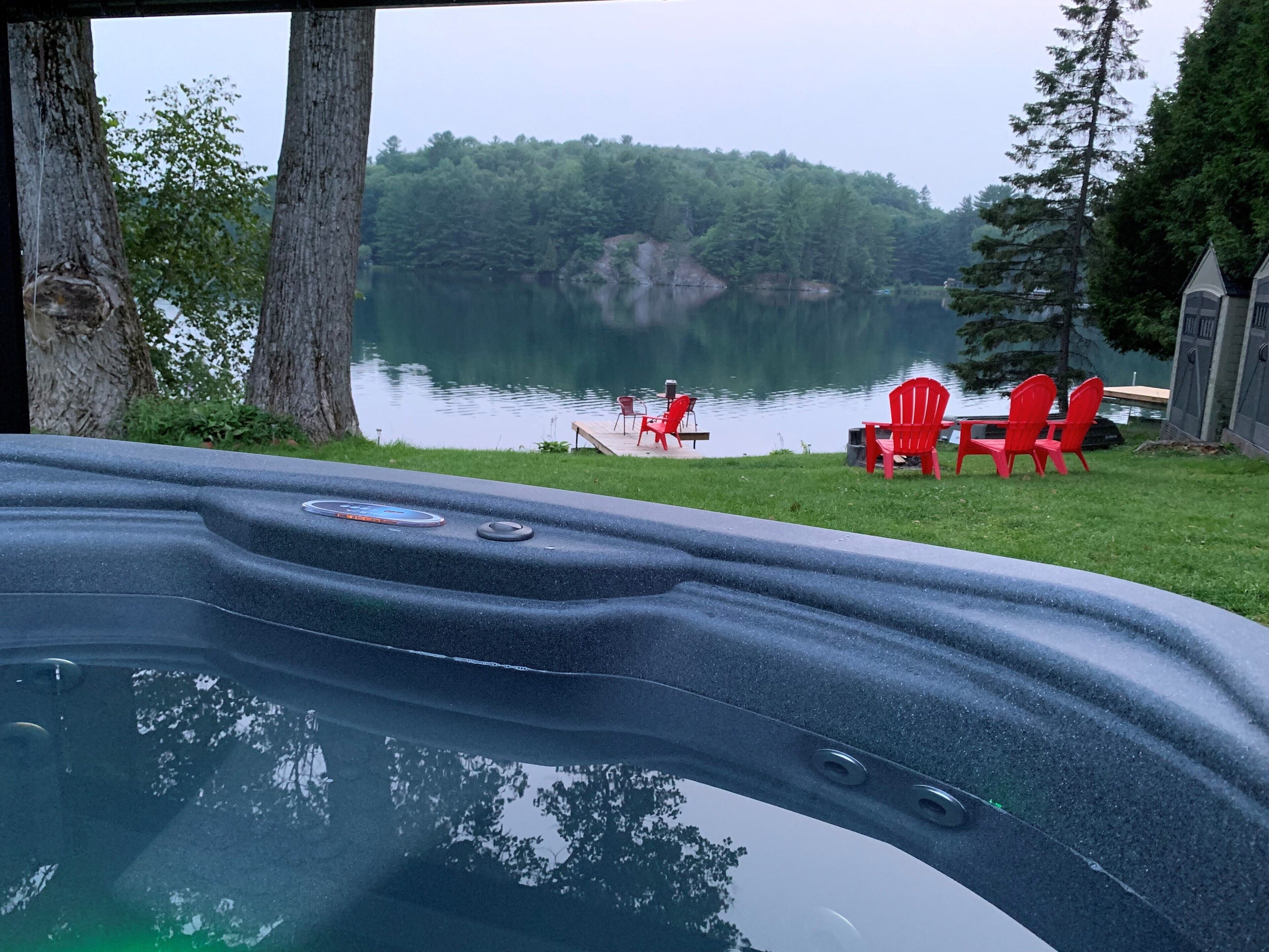 A hot tub with a view of a lake and forest. There are red chairs and a dock with a person fishing.