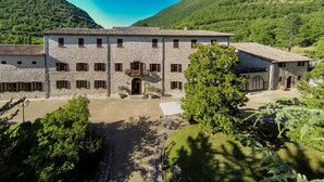 Ein steinernes Gebäude mit zahlreichen Fenstern und einem zentralen Eingang, umgeben von Bäumen und einer hügeligen Landschaft.