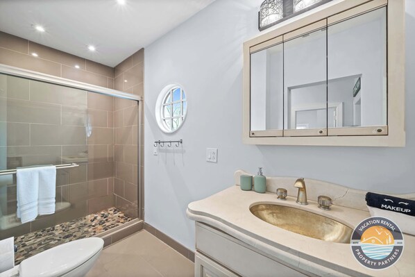 A bathroom with a glass shower enclosure, a round mirror, a beige sink, and a toilet.