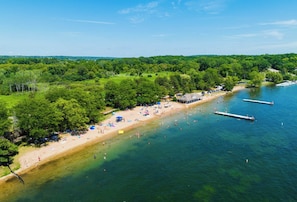 An aerial view of a beach with clear water, a sandy shore, and a forested area. - Lakeside Family Home  Walk to Beach (Williams Bay)