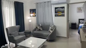 Una sala moderna con un sofá gris, una mesa de centro blanca y un sillón gris. Hay dos cuadros en la pared, una lámpara de pie y una puerta que da a otra habitación.