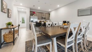 A modern dining area with a large wooden table, white chairs, and a kitchen in the background. - The Serenity Townhome (Kissimmee)