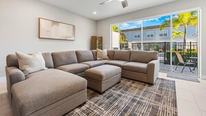 A modern living room with a sectional sofa, a ceiling fan, and a large window offering a view of a residential area. - The Serenity Townhome (Kissimmee)