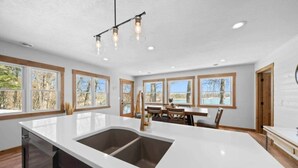 A spacious kitchen with a large island, wooden cabinets, and a view of a lake through large windows. - Mallard Lake Retreat (Jackson)