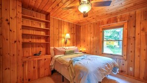 Une chambre dans une cabane en bois, avec un lit, des étagères, une fenêtre et un ventilateur de plafond.