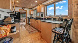 A modern kitchen with wooden cabinets, a black countertop, and a large window offering a view of the sky. - Buckfield Lakehouse | Private waterfront on Rangeley Lake (Rangeley)