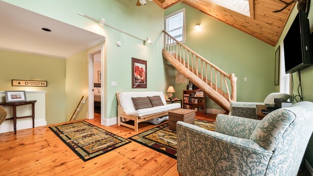 A cozy living room with a wooden floor, a sofa, a patterned armchair, a staircase, and a TV mounted on the wall. - Downtown Crossing Condo | Walk to Town | Pet Friendly | Close to Saddleback (Rangeley)