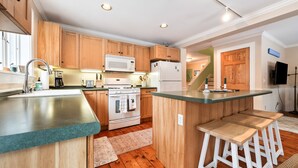 A modern kitchen with wooden cabinets, a central island with stools, a microwave, a refrigerator, and a stove. - Downtown Crossing Condo | Walk to Town | Pet Friendly | Close to Saddleback (Rangeley)