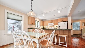 A modern kitchen with wooden cabinets, a central island with stools, a glass-top table, and a view of the outside. - Downtown Crossing Condo | Walk to Town | Pet Friendly | Close to Saddleback (Rangeley)