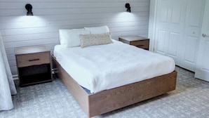 Quarto com cama de madeira, roupa de cama branca, duas mesas de cabeceira e luzes fixadas na parede.