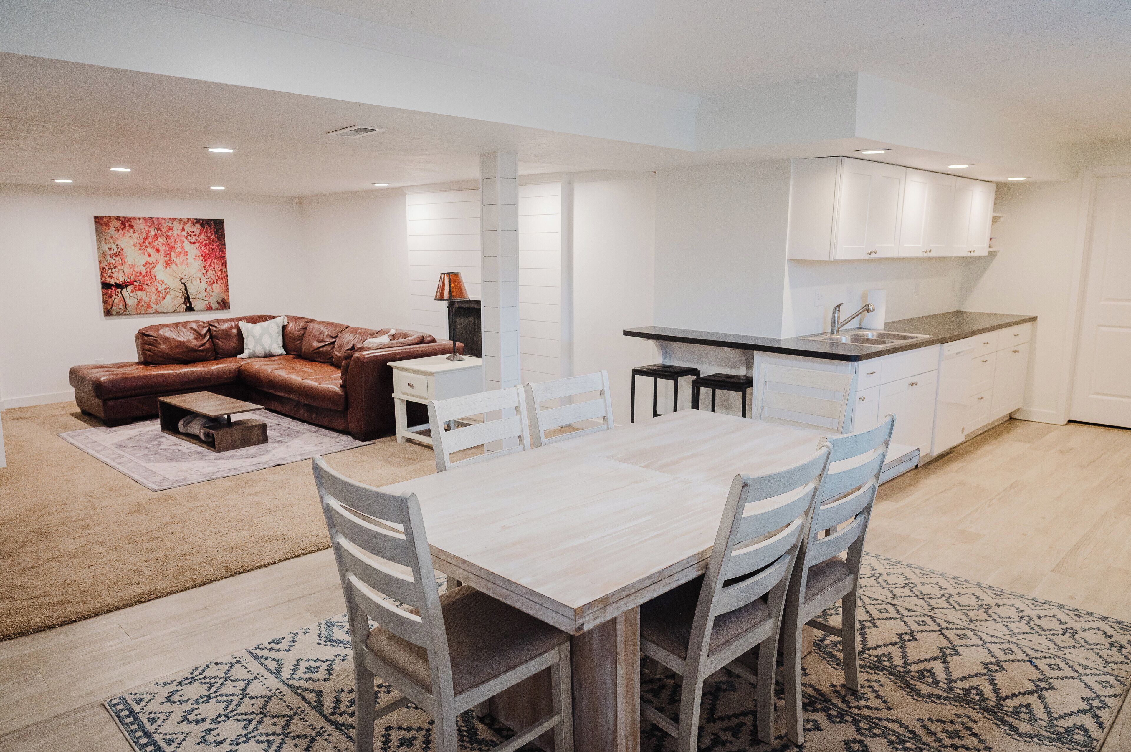 A modern apartment with a dining area, a brown leather sofa, a white dining table with four chairs, and a kitchen with white cabinets.