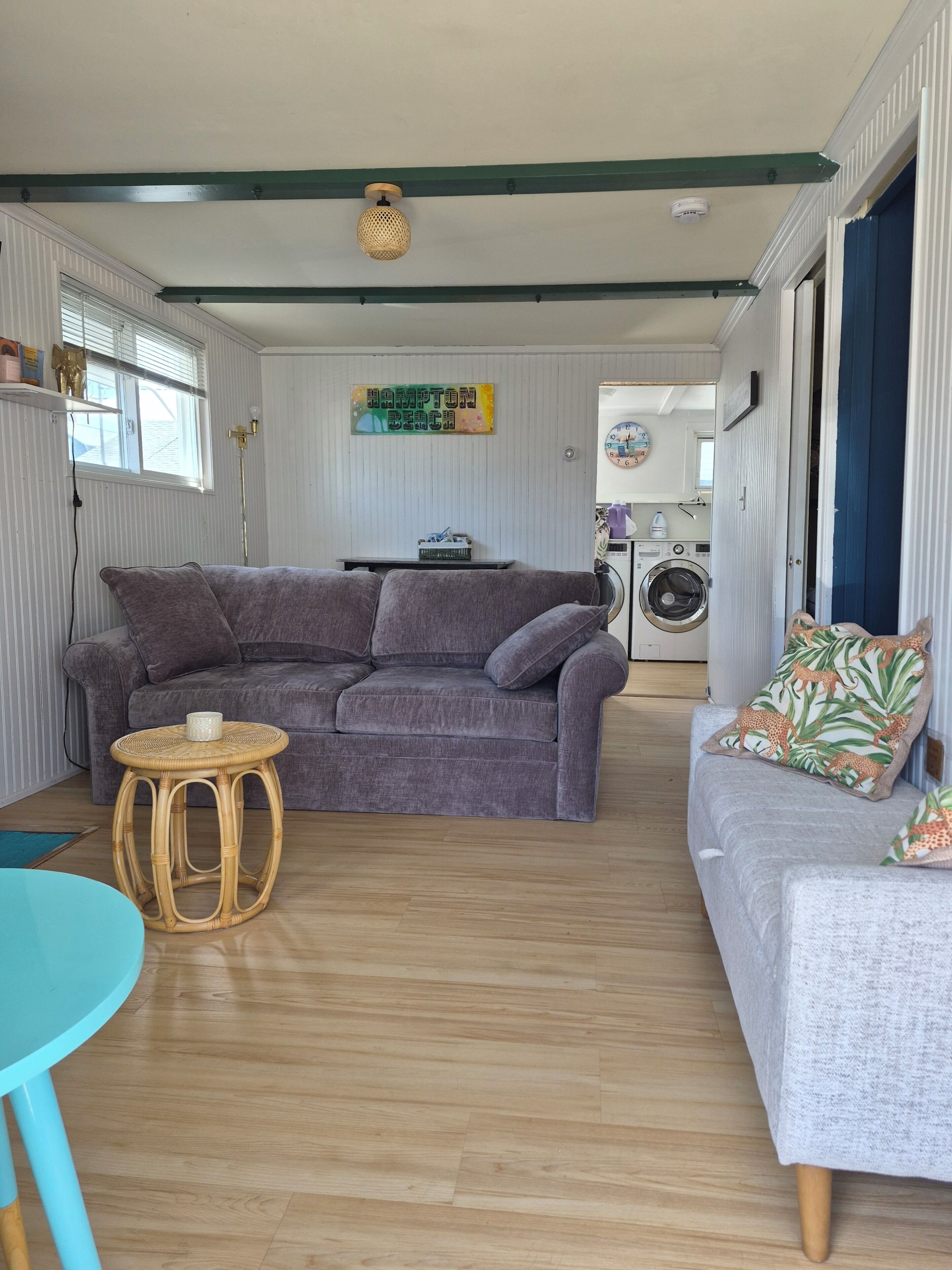 Een woonkamer met een grijze bank, een rond rieten bijzettafeltje en een grijze fauteuil met gekleurde kussens. Een wasmachine is zichtbaar op de achtergrond.