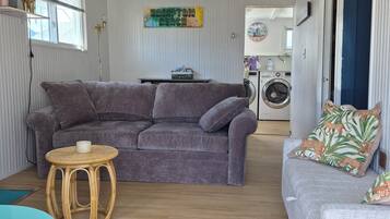 Een woonkamer met een grijze bank, een rond rieten bijzettafeltje en een grijze fauteuil met gekleurde kussens. Een wasmachine is zichtbaar op de achtergrond.