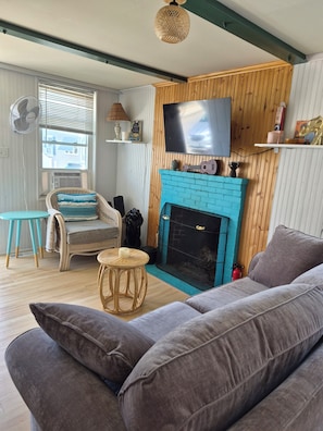 Una sala de estar con chimenea, un sofá, una silla, un televisor y un ventilador.