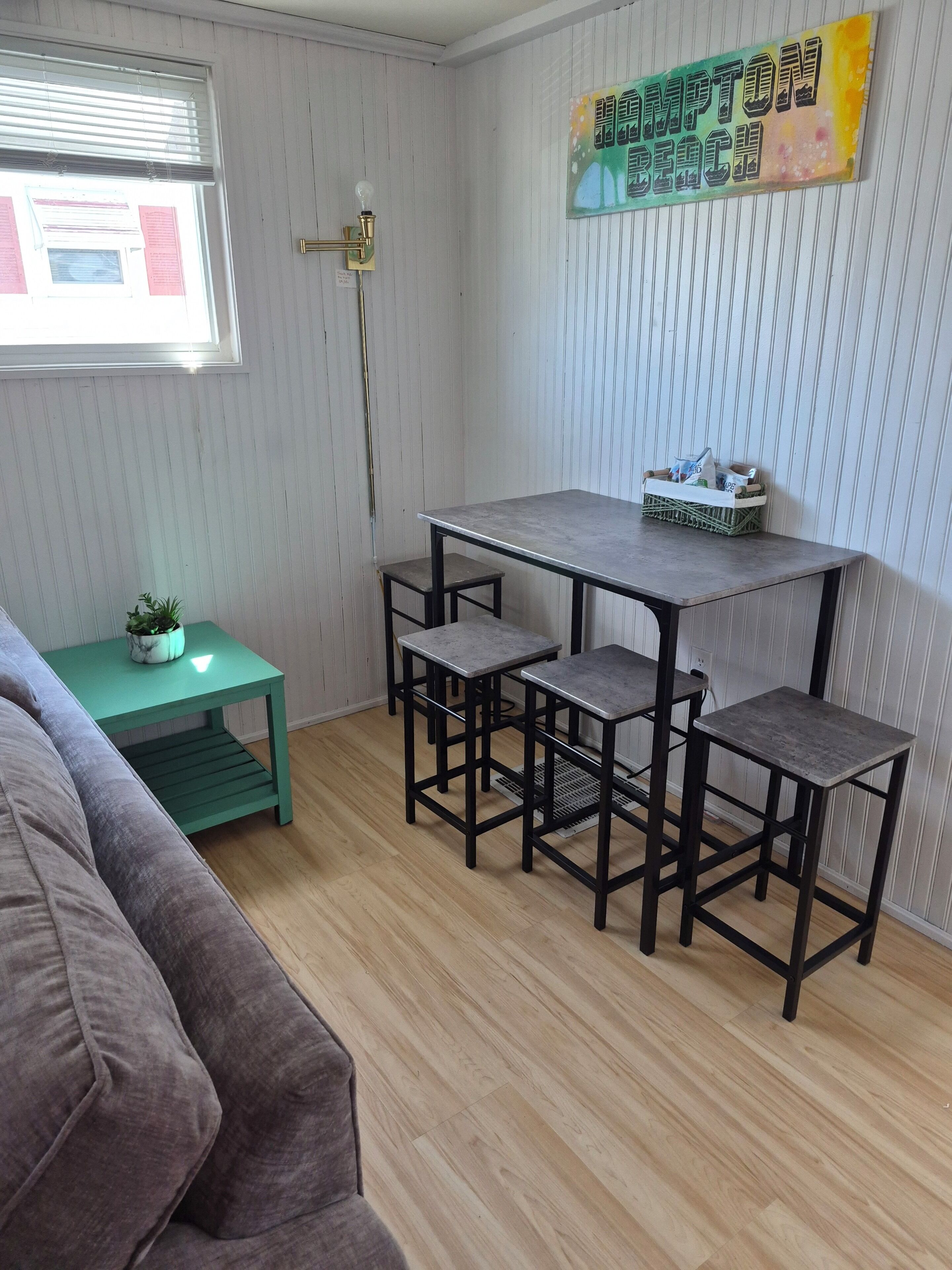 Ett rum med ett matbord och stolar, ett litet grönt sidobord med en växt och ett fönster med persienner.