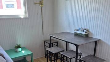 Ett rum med ett matbord och stolar, ett litet grönt sidobord med en växt och ett fönster med persienner.