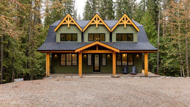 Une cabane en rondins à deux étages, dotée d’un porche et d’une allée en gravier, entourée d’une forêt dense.
