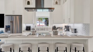 A modern kitchen with white cabinetry, a stainless steel refrigerator, a large island with bar stools, and a view of the outdoors. - Live in Luxury at This Brand New Tybee Island Escape | Heated Pool Access (Tybee Island)