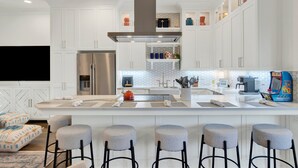 Uma cozinha moderna com armários brancos, uma ilha central com banquetas, uma geladeira de aço inoxidável e uma cafeteira.
