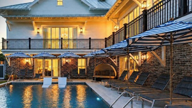 Área da piscina com espreguiçadeiras e guarda-sóis, parede de tijolos e varanda iluminada.