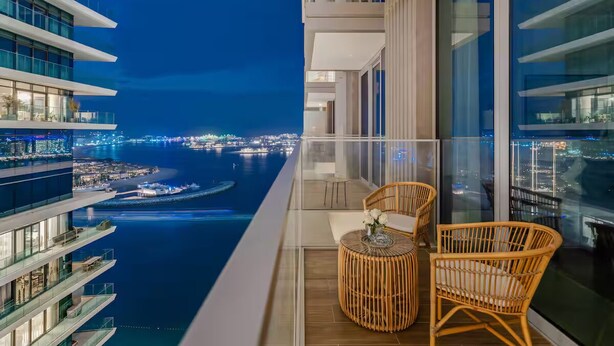 Un balcon avec des fauteuils et une table en osier, offrant une vue sur le paysage urbain et l’eau la nuit.