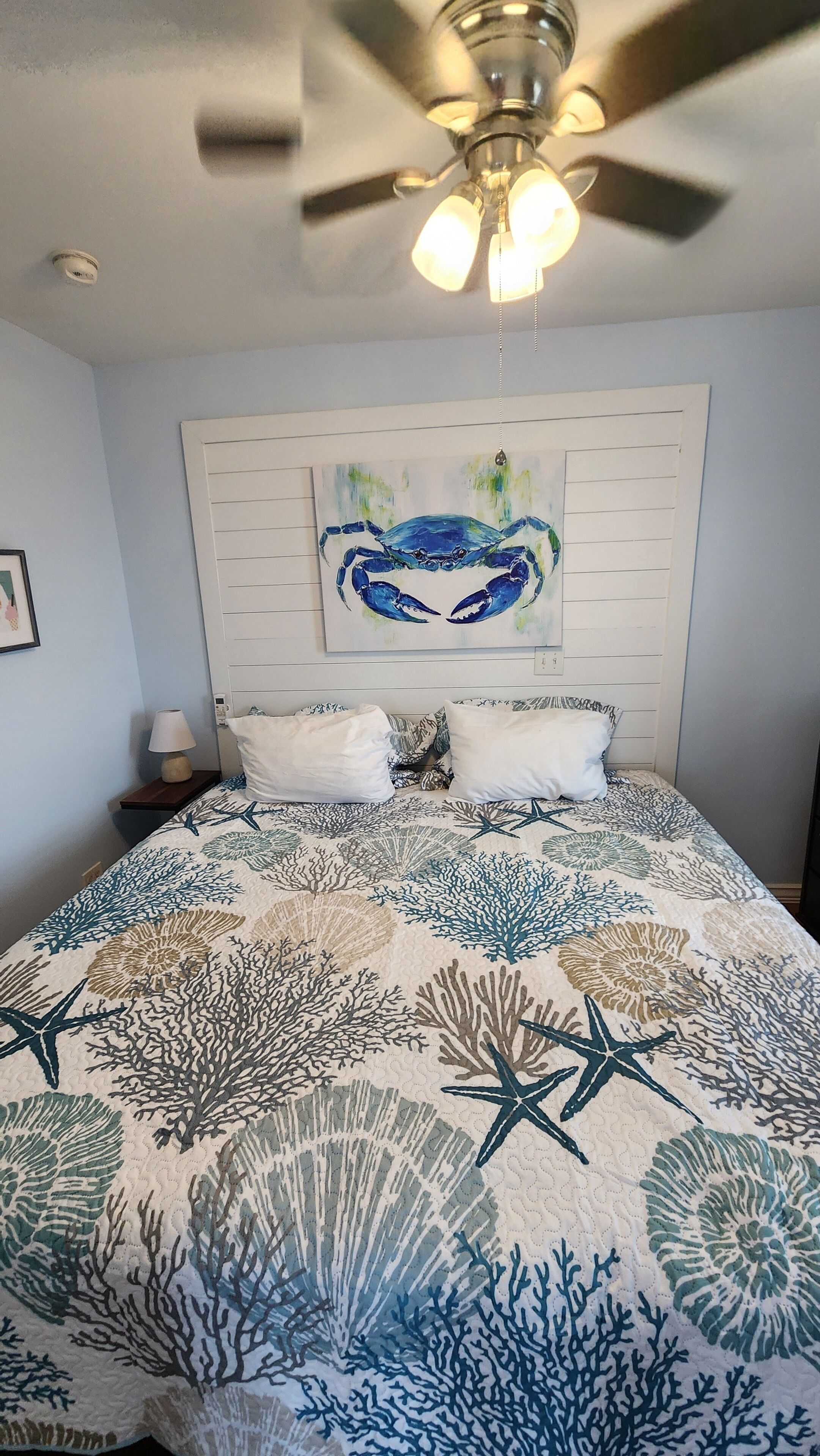 Une chambre à coucher avec un lit, un ventilateur de plafond, un tableau représentant un crabe et une table de chevet avec une lampe.