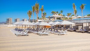 Une plage bordée de rangées de chaises longues et de parasols, avec des palmiers et des bâtiments modernes en arrière-plan.