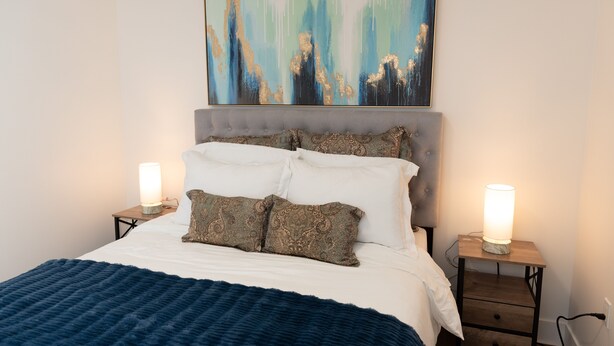 Una cama bien hecha con una colcha azul, dos lámparas de noche y un cuadro abstracto grande sobre la cama.