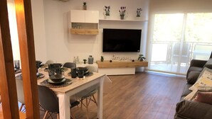 A modern living room with a dining area, a flat-screen TV mounted on the wall, and a large window providing natural light. - RIERA Modern Apartment in the Heart of Roses (Roses)