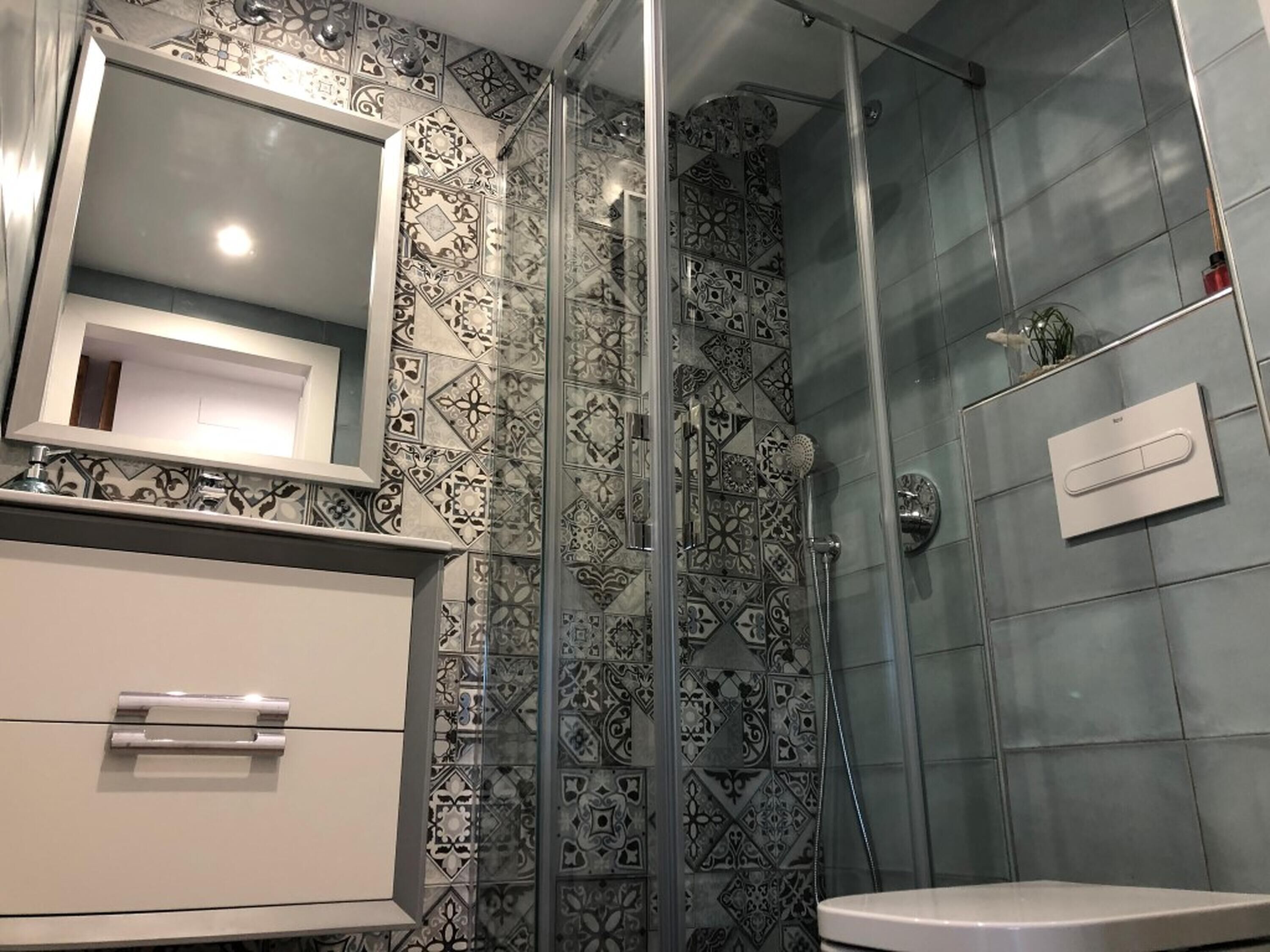 Um banheiro com azulejos estampados, box de vidro, pia branca e espelho.