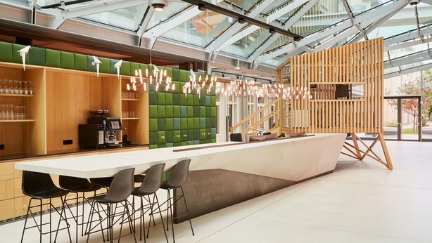 Een moderne bar met een lange toonbank, zwarte stoelen en een zithoek met groene kussens.