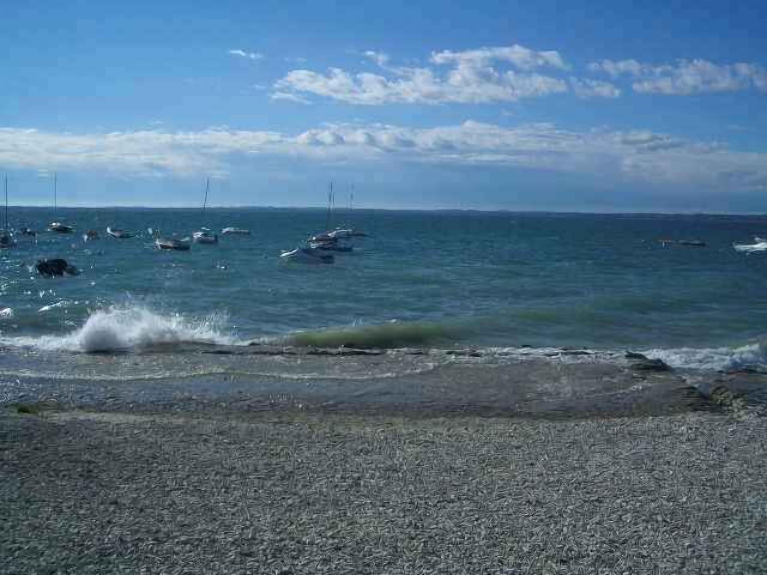 Ein Kiesstrand mit Booten im Wasser und einem wolkenlosen Himmel.