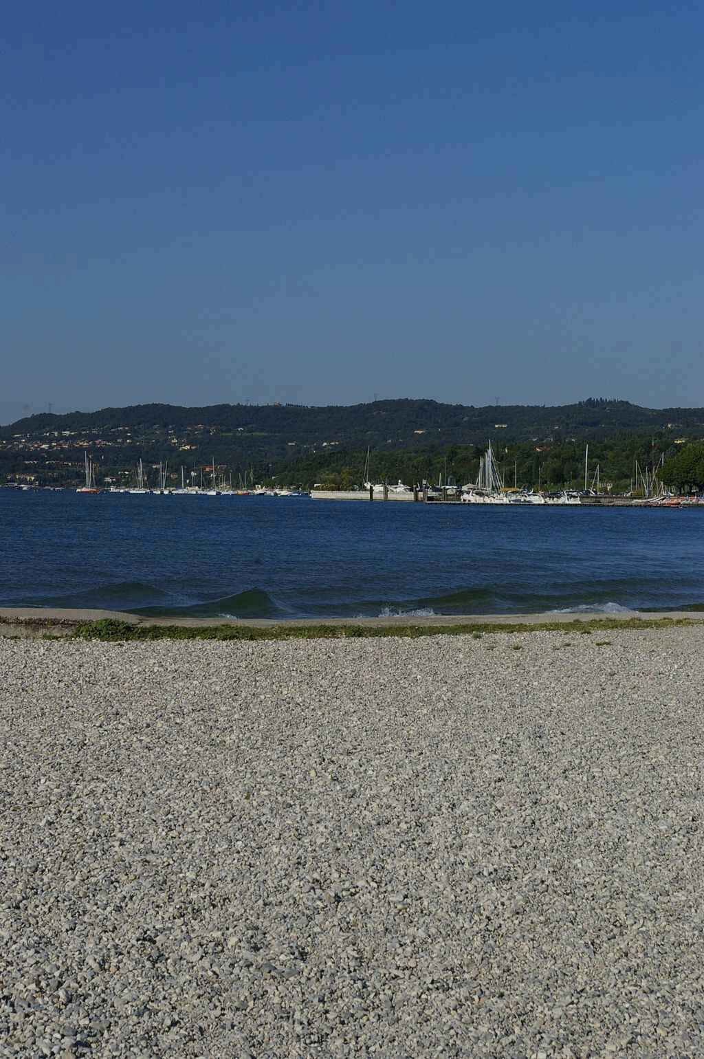 Ein Kiesstrand mit im Hintergrund vor Anker liegenden Booten.