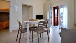 A room with a dining table, four chairs, a TV, and a bed with orange bedding. - Holiday apartment with air conditioning and balcony (Bibione)