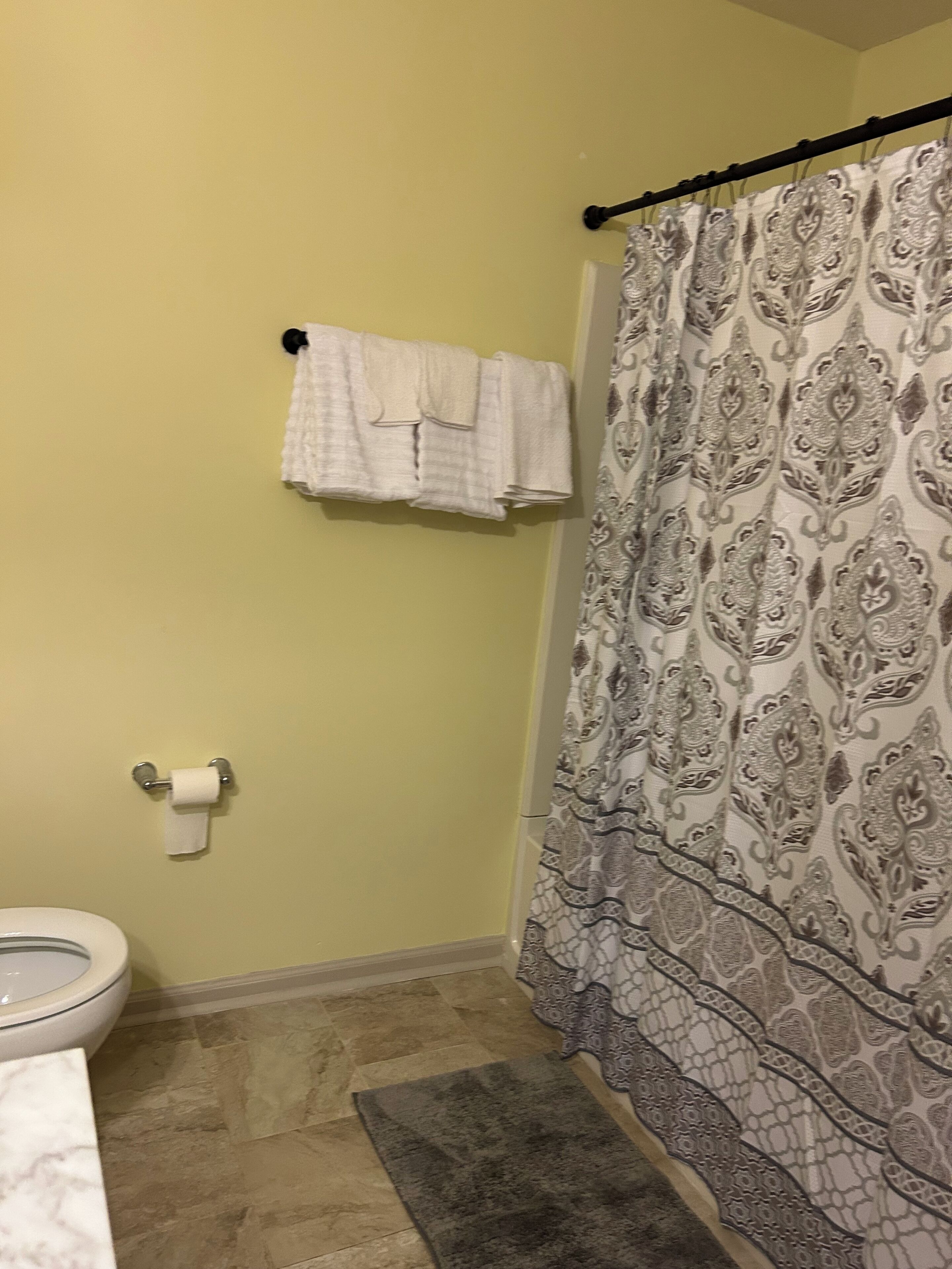 A bathroom with a toilet, a towel rack with folded towels, a patterned shower curtain, and a small rug on the floor.
