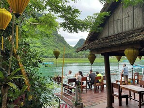A riverside dining area with wooden tables and chairs, surrounded by lush greenery and traditional lanterns. - Phong Nha Coco House Homestay  (Phong Nha)