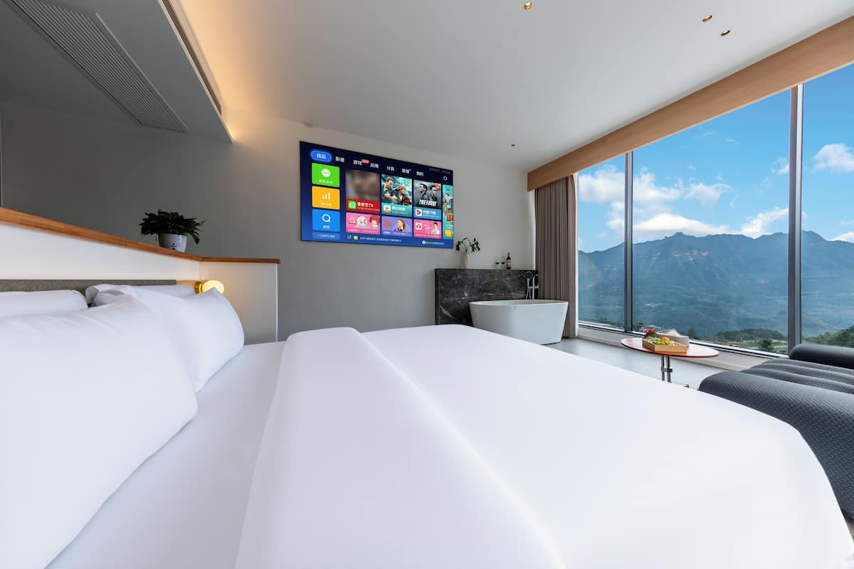 Une chambre d’hôtel moderne avec un grand lit, un téléviseur fixé au mur qui affiche différentes applications, une petite table avec une plante et une vue sur les montagnes par de grandes fenêtres.