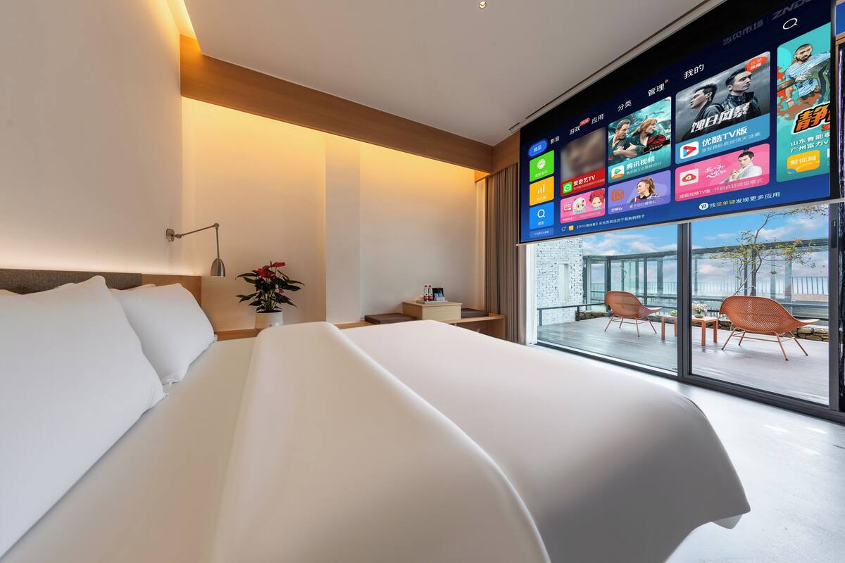 Une chambre d’hôtel moderne avec un grand lit, un bureau avec une lampe, une petite table avec une plante et une grande fenêtre donnant sur un balcon équipé de chaises.