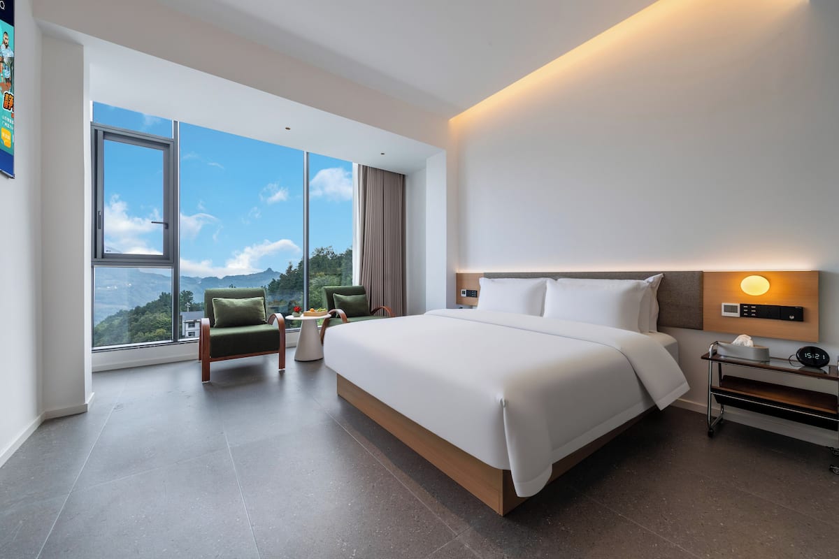 Une chambre d’hôtel moderne dotée d’un grand lit, d’un coin salon avec un fauteuil et une petite table, d’une table de chevet avec une lampe, et offrant une vue sur l’extérieur grâce à de grandes fenêtres.