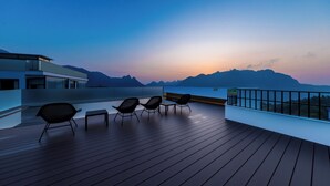 Terraza de madera con cuatro sillas y una mesa, con vista a un paisaje montañoso al atardecer.