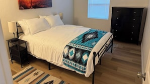 Quarto com cama, mesas de cabeceira, um criado-mudo, janela com persianas, uma obra de arte na parede e um armário.