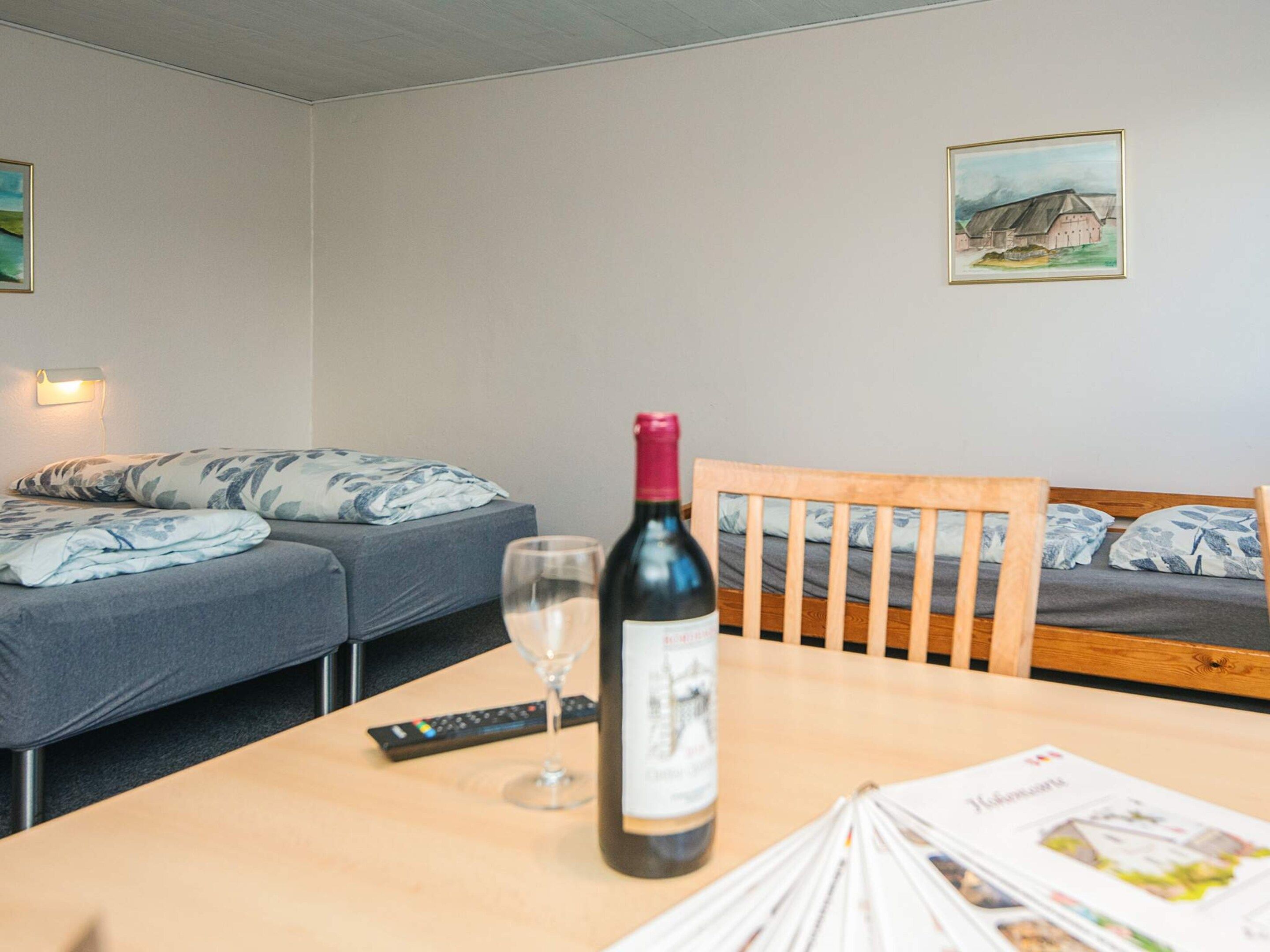 A room with a bed, a table with a bottle of wine, a glass, and a remote control.