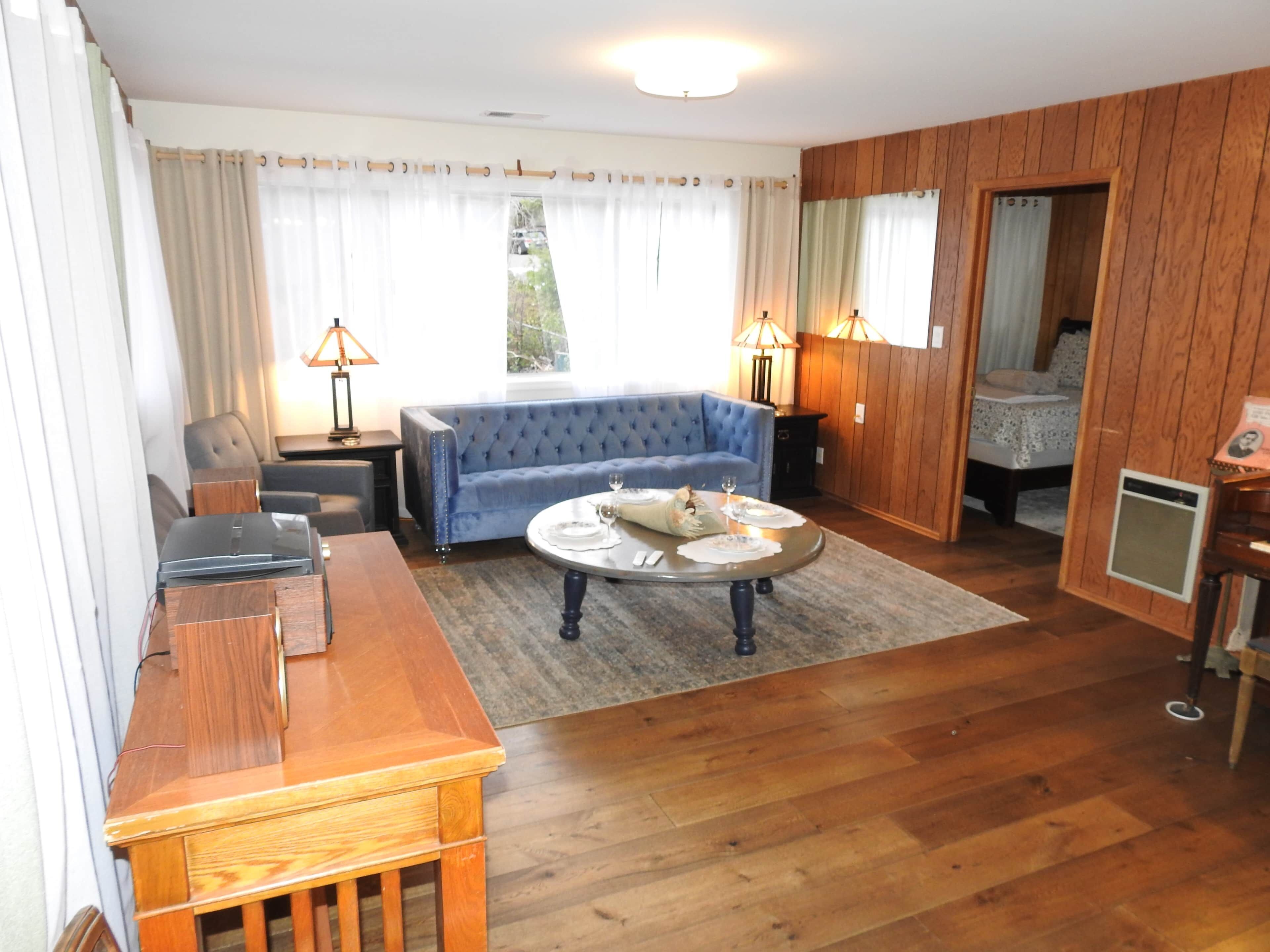 Una habitación con un sofá azul, una mesa de centro de madera, una mesita con lámparas y una cama con cabecero de madera.